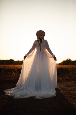 Doğada günbatımında arkası dönük mutlu bir kadın. Kafasında şapkası olan uzun beyaz elbiseli bir kadın.