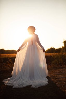 Doğada günbatımında arkası dönük mutlu bir kadın. Kafasında şapkası olan uzun beyaz elbiseli bir kadın.