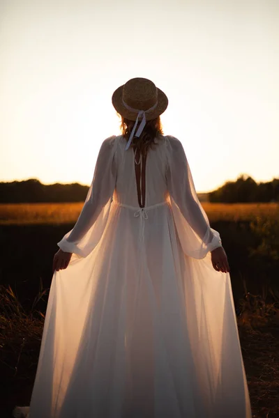 快乐的女人 背对着日落站在大自然中 头戴帽子 身穿白色长裙的女人 免版税图库照片