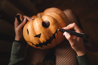 Tatlı genç bir kız oturma odasında bir masada oturuyor Cadılar Bayramı için yüzünü büyük bir balkabağına boyuyor. Cadılar Bayramı tatili ve aile hayatı.