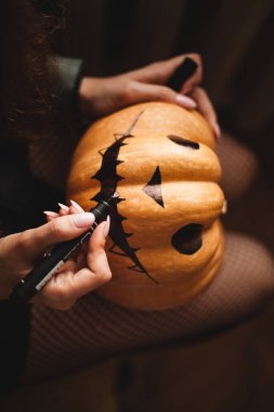 Tatlı genç bir kız oturma odasında bir masada oturuyor Cadılar Bayramı için yüzünü büyük bir balkabağına boyuyor. Cadılar Bayramı tatili ve aile hayatı.