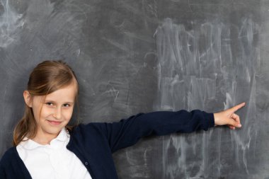 A girl stands smiling at a chalkboard and points to an empty space ready for writing with chalk. clipart