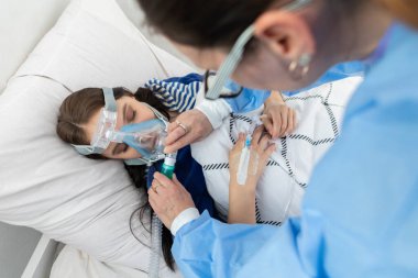 Doktor vantilatörün bağlantısını kontrol ediyor. Genç bir hastane yatağında yatıyor. Solunum yolundaki oksijen maskesi..