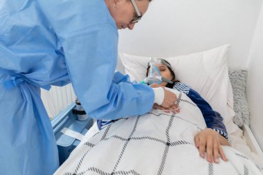 Doktor vantilatörün bağlantısını kontrol ediyor. Genç bir hastane yatağında yatıyor. Solunum yolundaki oksijen maskesi..