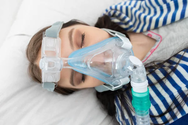 Oxygen mask attached to the respiratory tract. The teenager lies in a hospital bed. A seriously ill young girl.