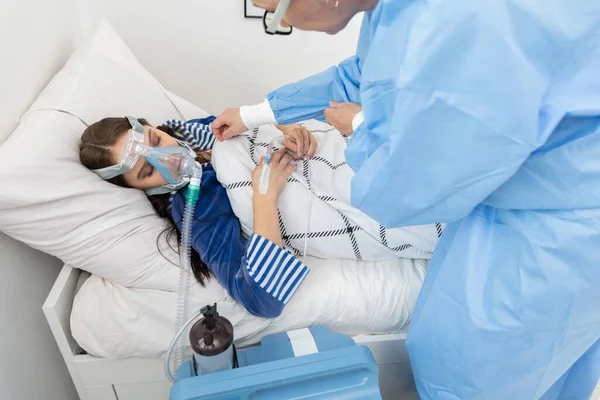 The doctor controls the connection of the ventilator. The teenager lies on a hospital bed. Oxygen mask on the respiratory tract.