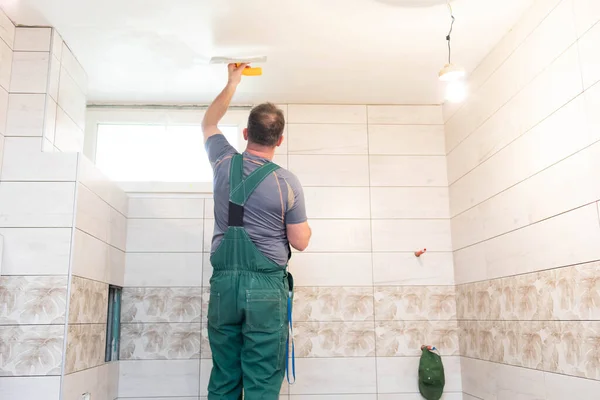 The worker applies plaster on the ceiling of the renovated bathroom. Construction worker in middle age. Green work clothes.