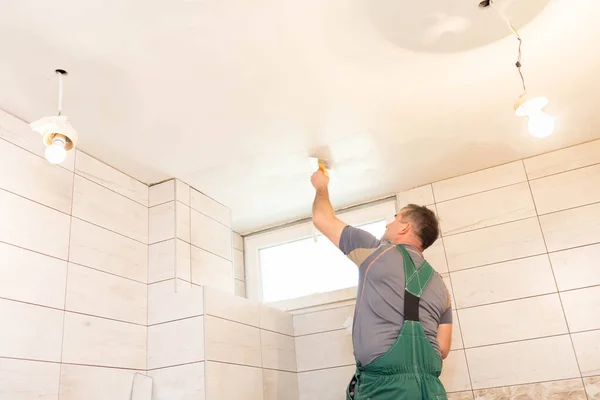 The worker applies plaster on the ceiling of the renovated bathroom. Construction worker in middle age. Green work clothes.