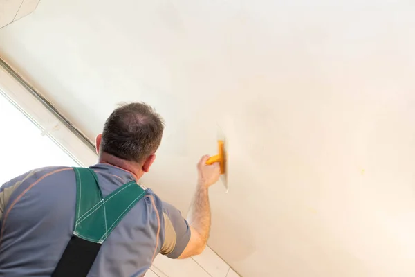 The worker applies plaster on the ceiling of the renovated bathroom. Construction worker in middle age. Green work clothes.