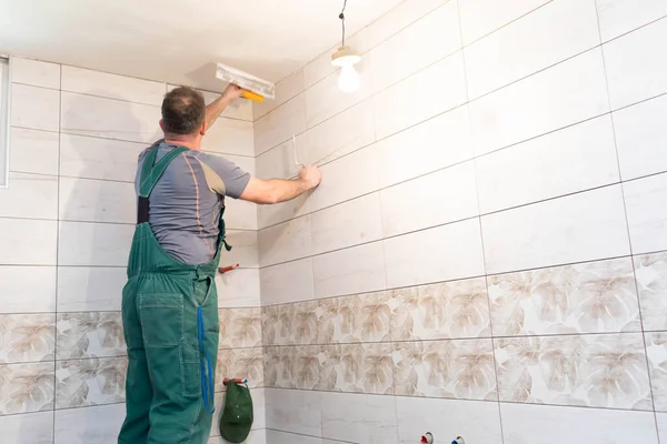 The worker applies plaster on the ceiling of the renovated bathroom. Construction worker in middle age. Green work clothes.