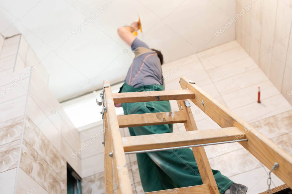 Applying gypsum finish on the ceiling of the renovated bathroom. Construction worker in middle age. Green work clothes.