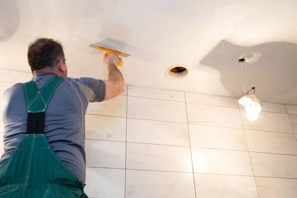 Un trabajador de la construcción aplica un compuesto de nivelación al techo con una paleta de acero inoxidable. Obras en altura desde el techo . — Foto de Stock