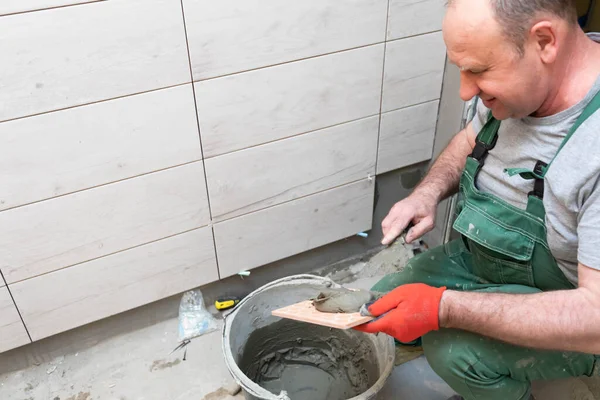 Un trabajador de la construcción aplica adhesivo de cemento a una baldosa de cerámica y lo extiende a fondo sobre toda la superficie . — Foto de Stock