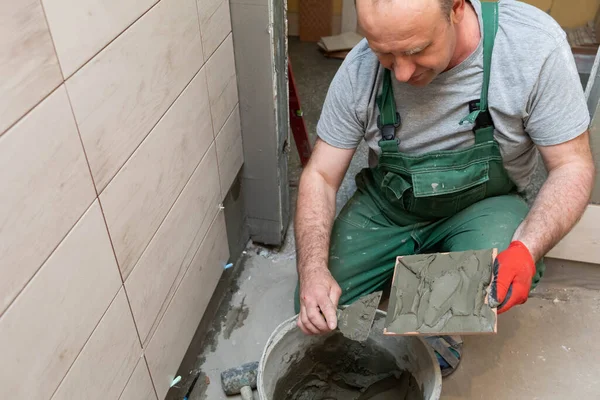 Un trabajador de la construcción aplica adhesivo de cemento a una baldosa de cerámica y lo extiende a fondo sobre toda la superficie . — Foto de Stock