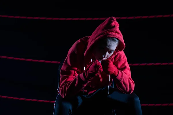 A lost fight of a strong MMA player. He is sitting in a chair. Right after losing the fight. Sad woman after a fight in the ring. In a hoodie.