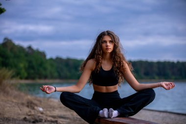 Huzurlu ve sakin bir sahne. Suların kenarında huzur içinde meditasyon yapan bir kadın, bereketli doğayla çevrili.