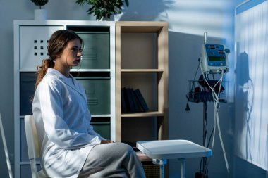 Bir kadın düşünceli bir şekilde klinik bir ortamda oturuyor, sakin, sakin ve odaklanmış bir atmosfere yansıyor.