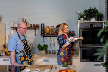 Neşeli bir çift modern bir mutfakta birlikte yemek pişiriyor, canlı önlükler giyiyor ve taze malzemeler kullanıyor.