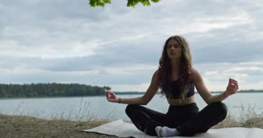 Huzur içinde oturan bir kadın, güzel ve huzurlu doğayla çevrili sakin bir göl kenarında meditasyon yapıyor.