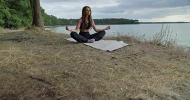 Bir kadın huzur içinde yoga meditasyonu yapıyor. Huzurlu bir göl kenarında. Etrafı güzelleşen doğayla çevrili.