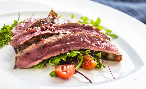 Close-up portion of juicy flank steak from vegetable salad salt pepper and rosemary on white plate.