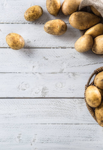Ripe potatoes in burlap sack and wooden basket freely lying on wooden board.