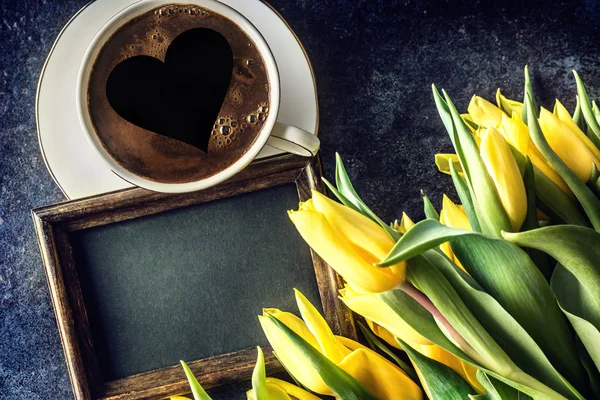 Top of view cup form coffee with heart empty chalkboard and spring tulips on wooden table.