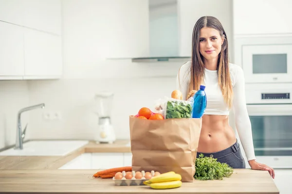 Jovem mulher de pé na cozinha com compra na mesa — Fotografia de Stock