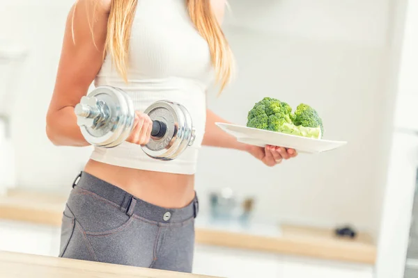 Mujer joven con figura deportiva en la cocina sosteniendo pesas —  Fotos de Stock