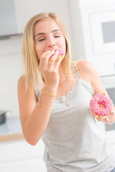 Young blonde girl eats pink donuts in home kitchen with taste em — Stock Photo, Image