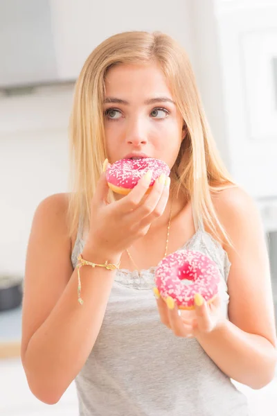 Joven chica rubia come rosados donuts en casa cocina con gusto em —  Fotos de Stock