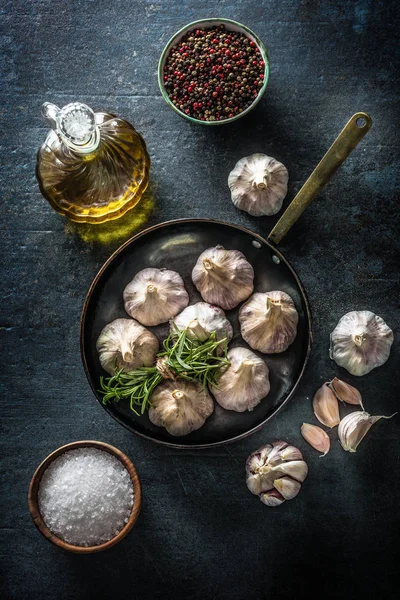 Garlic cloves and bulb in pan with fresh rosemary olive oil salt and spice on concrete table