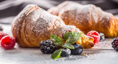 Taze çıtır kruvasanlar üzerine ahududu böğürtlen ve yaban mersini serpiştirilmiş şeker tozu serpiştirilmiş.