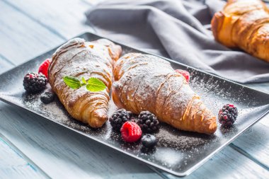 Taze çıtır kruvasanlar üzerine ahududu böğürtlen ve yaban mersini serpiştirilmiş şeker tozu serpiştirilmiş.