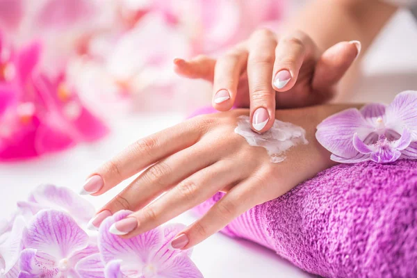 Hermosas manos femeninas aplicando crema de manos. Toalla púrpura y decoración de orquídeas. Concepto saludable - Cuidado y bienestar de la piel del spa de uñas —  Fotos de Stock