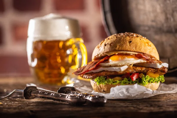 Hamburger with chicken, fried egg and bacon, salad, peppers with a rustic knife and barrel on the side along with a draft beer