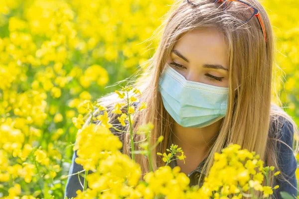 アレルギーブロンド女の子でA保護マスク見ますザ黄色の花座っている上の牧草地 — ストック写真
