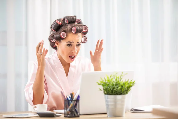 Upset woman with hair rollers and night dress looks at laptop screen with a shocked expression.