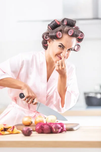Woman in nightdress and hair rollers holds her nose from a smelly onion that she cuts on a cutting board.