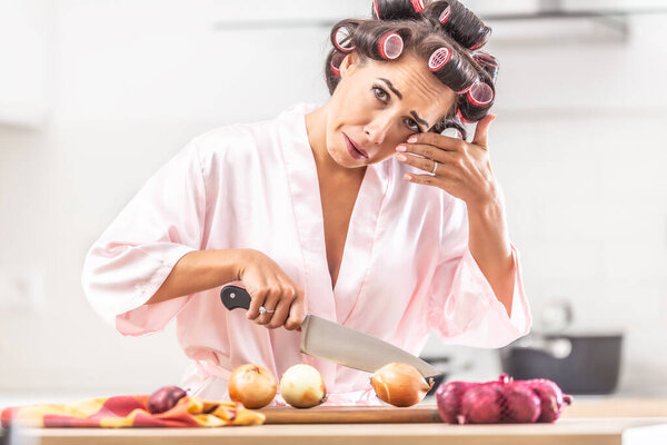 Woman in hair curlers and nighgown cries from onion cutting in the kitchen, wiping the tear from her eye.