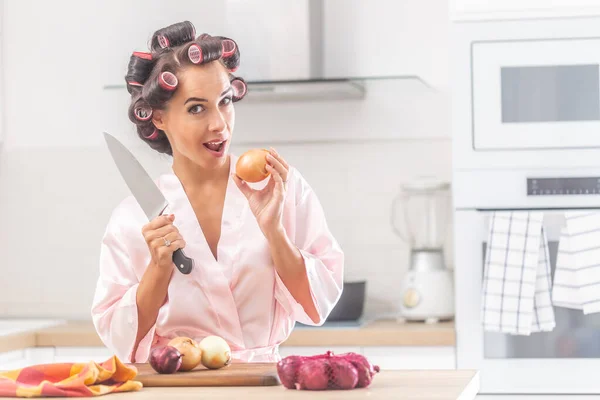 漂亮的女人拿着刀和洋葱站在厨房里 穿着卷发和睡衣 — 图库照片