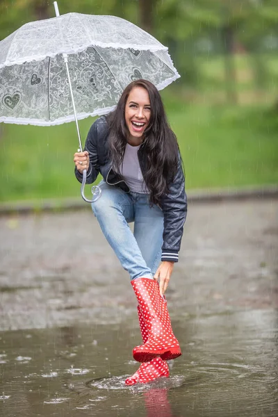 Woman laughing at water leaking to her rain boot, taking it off, while she keeps protecting herself againt the rain by an umbrella.