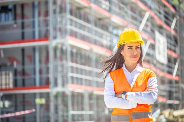 Female site supervisor wearing safety vest and helmet stands confidently in front of the construction covered in scaffolding.