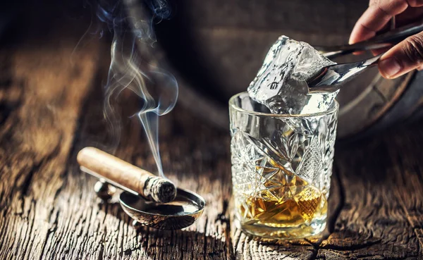 Ice cube being put into an ornamental cup of whisky placed on a old fashioned wood and burning cigar aside — Stock Photo, Image