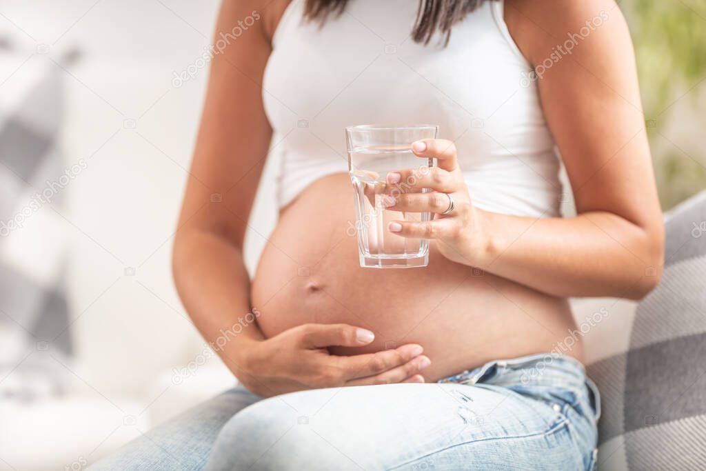 Pregnant female holding her belly with one hand holds a glass of water in the other hand.