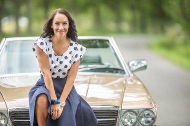 Muhteşem esmer, kameraya gülümsüyor. Teh Doğa 'da yol kenarında üstü açık bir arabanın ön kaputunda oturuyor..