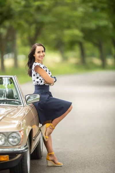 Beautiful woman in a skirt and high heels leans her back against a vintage convertible car parked on the roadside.