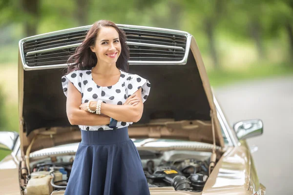Confident woman stand in front of a car with open bonnet, arms crossed, smiling into the camera.