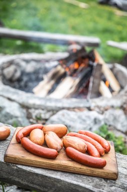 Arka planda kamp ateşinde yanmaya hazır ahşap kesme tahtasındaki sosisler.