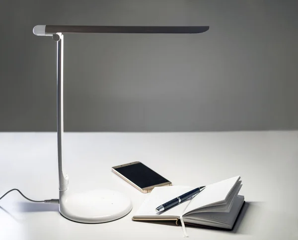 A smartphone and a diary with a pen on the table under the desktop LED lamp. Selective focus.